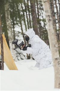 A white-clad person crouches in the snow with a paintball gun.