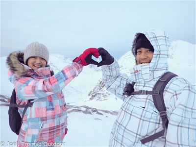 Two people in winter clothing forming a heart shape with their hands in snowy mountains.