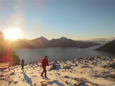To personer går i snødekt fjellterreng med solnedgang og vann i bakgrunnen.