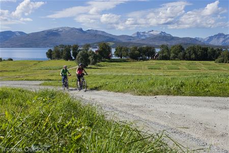 Two people cycling on a gravel path through grassy fields, with mountains, water, and trees in the background.