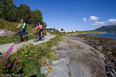 Two people cycling on a coastal path surrounded by flowers and sea, with mountains and houses in the background.