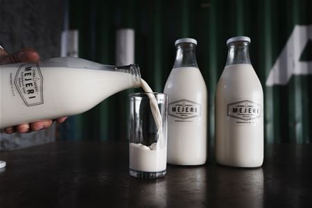 Någon häller upp mjölk från en glasflaska i ett glas, brevid står två fulla glasflaskor med mjölk.