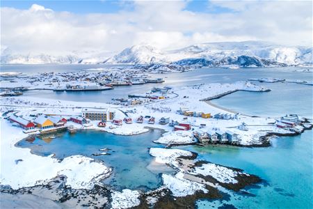 Fargerik kystlandsby med snø, fjell og sjø under delvis skyet himmel