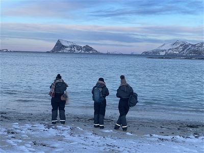 Tre personer på snødekt strand med fjord og fjell i bakgrunnen under blårosa himmel