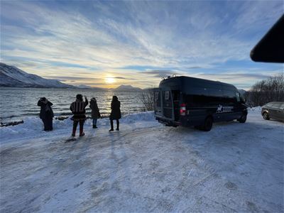 Folk ved snødekt utsiktspunkt med fjord, fjell og solnedgang