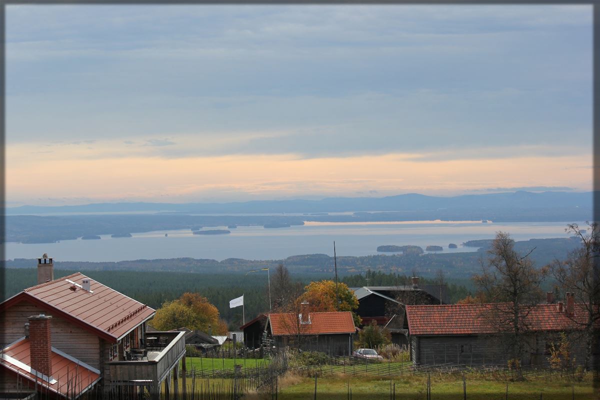 Vy över Fryksås med gråa fäbodstugor i förgrunden och Orsasjön i bakgrunden.