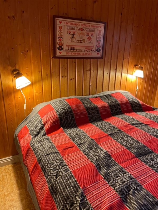 Double bed with grey and red bedspread against a pine wall. 