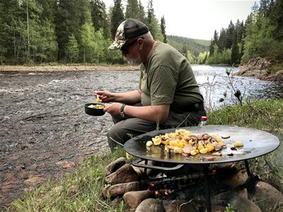 En man sitter och äter bredvid älven med en murikka bredvid som steker maten.