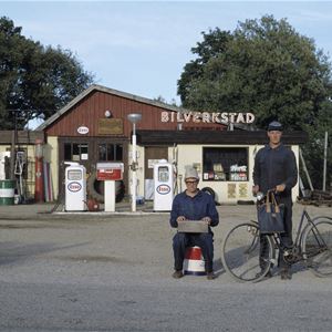 Två män i blå overaller en med en cykel, den andra sitter ner, i bakgrunden en bensinmack.