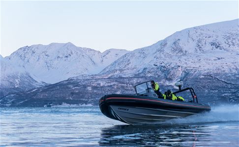 En RIB-båt kjører fort i fjorden med snødekte fjell i bakgrunnen