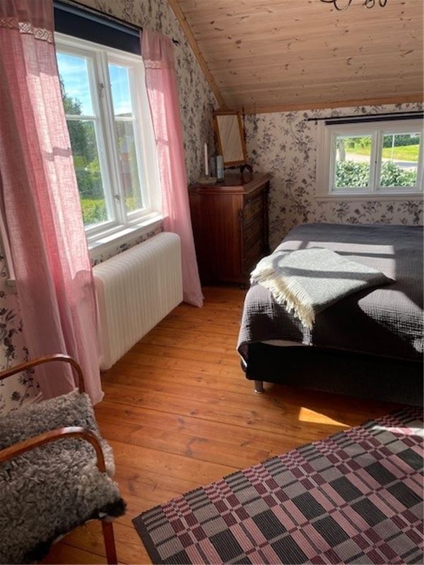 Ett sovrum med två sängar, blommiga tapeter, fårskinnsfåtölj, rosa gardiner.