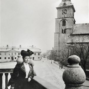 En svartvit bild på en kvinna i svart hatt som står lutad mot ett stenräcke, Kristine kyrka i bakgrunden.