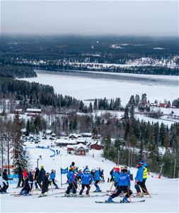 Många slalomåkare står långt upp i backen med utsikt över by och sjö.