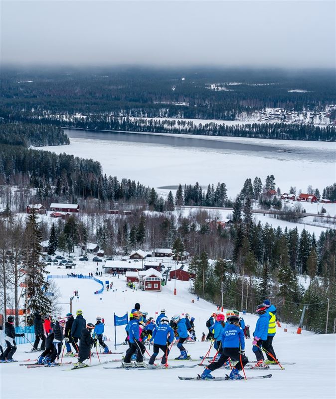 Many slalom skiers stand far up the hill with a view of the village and lake.