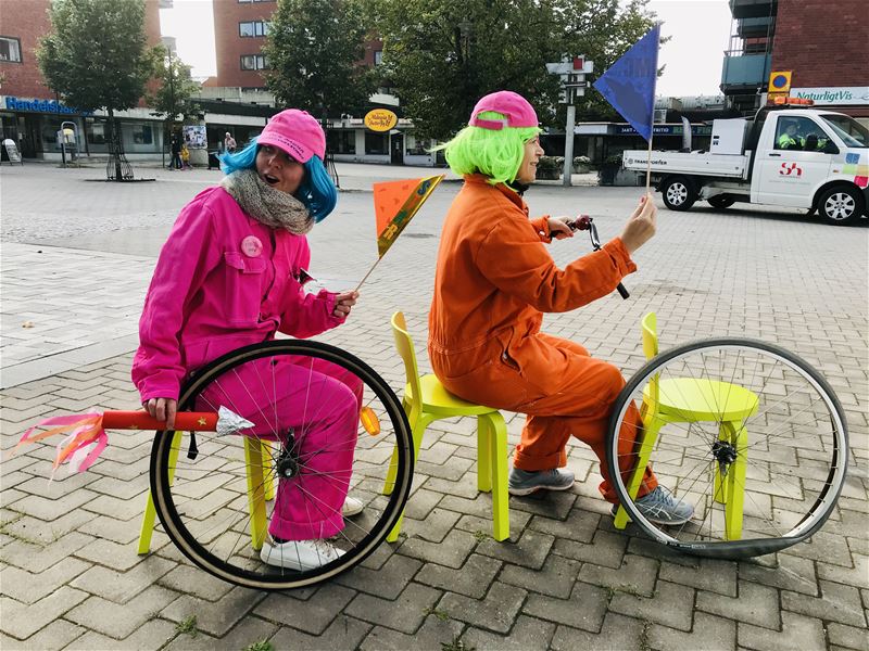 Två personer med färgglada overaller och kepsar leker cykel med pallar och cykelhjul.