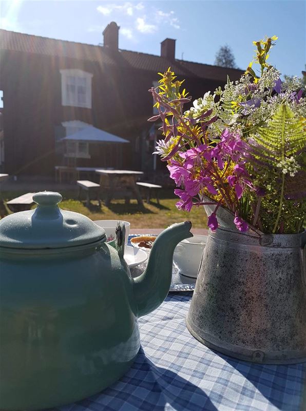 Dukat bord utomhus med kaffekanna och blommor i vas. blå och vitrutig duk med rött timrat hus i bakgrunden.