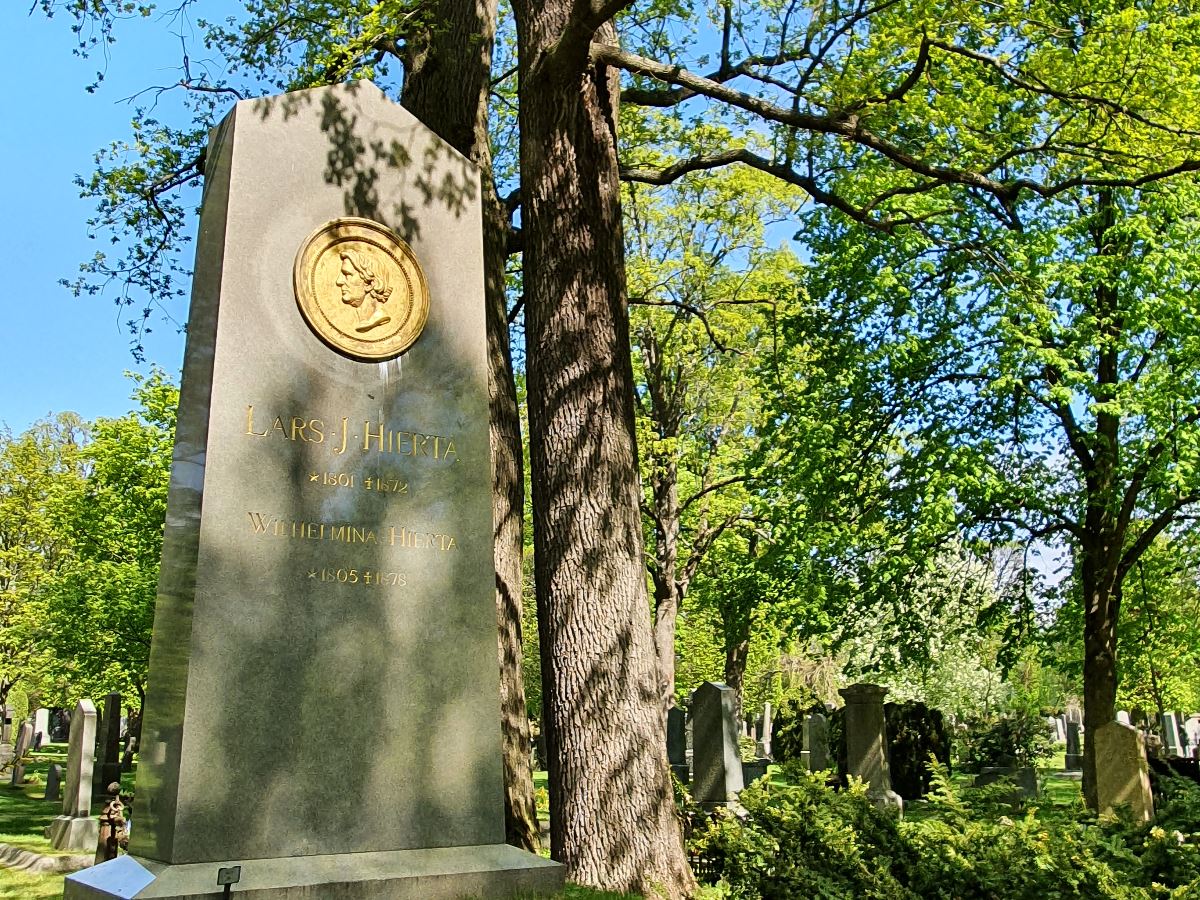 Stor och hög gravsten i en grön dunge med buskar och träd och en blå himmel 