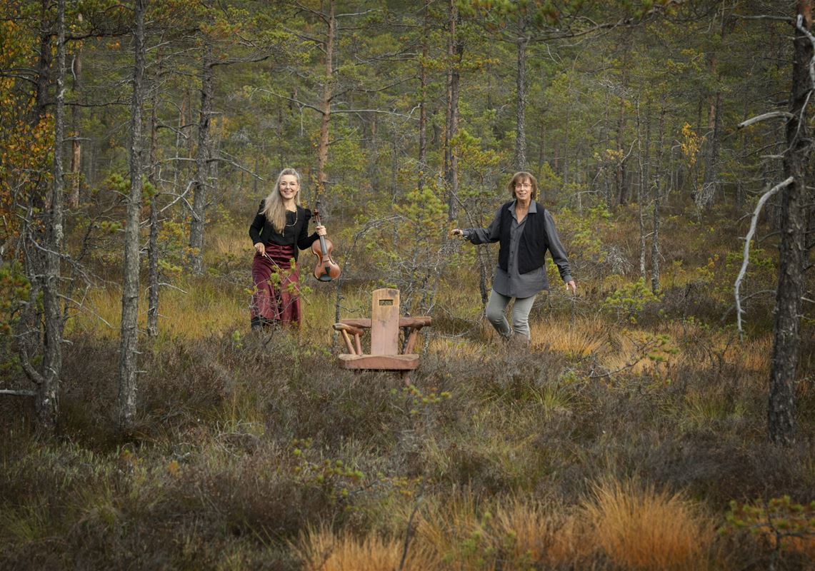 Musikerna Maria Sedell och Livet Nord kommer vandrande i skogsmark där en stol står mitt i skogen.