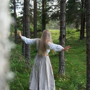 En ljusklädd kvinna går från kameran med utsträckta armar bland trädstammar. 