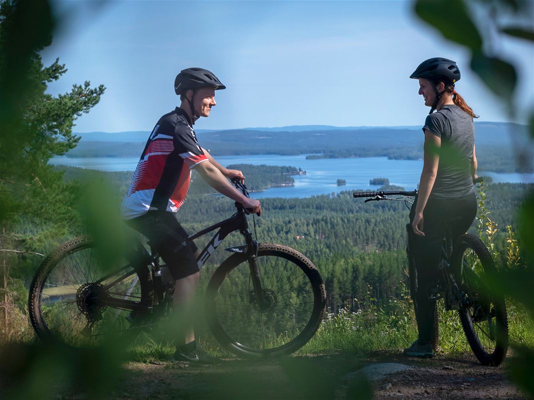 En man och en kvinna med varsin cykel, tittar på varandra, vacker utsikt med vatten berg och skog i bakgrunden.