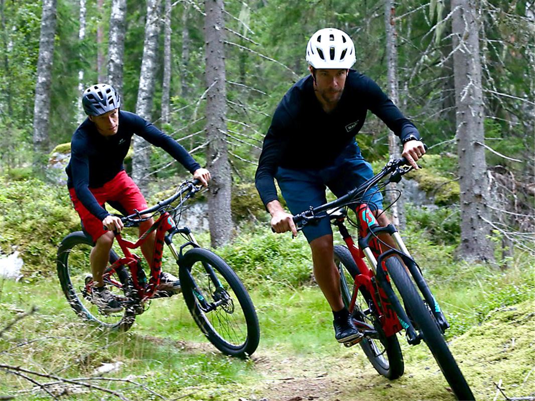 Två män som cyklar på en skogsstig, båda har var sin röd cykel.