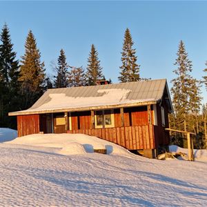 En röd stuga med veranda i snölandskap med höga granar bakom. 