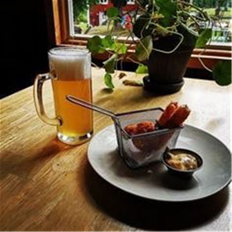 En öl i en sejdel, en tallrik med en nätkorg med rotsaker och en liten skål med dipp, en växt och ett fönster i bakgrunden.