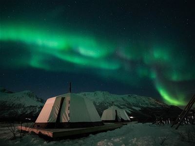 Northern lights over snow-covered mountains and tents on platforms during a winter night.