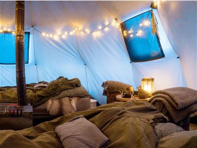 Tent interior with multiple beds, a wood-burning stove, string lights, and blue window coverings.