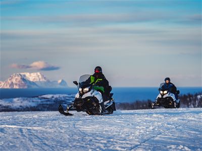 To personer på snøscooter i snødekt landskap med fjell og hav i bakgrunnen