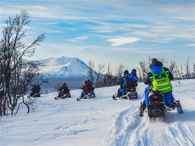 Gruppe på snøscootere i snødekt landskap med guide i gul vest, trær og snødekt fjell i bakgrunnen