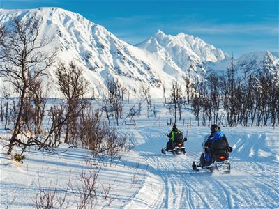 To personer på snøscooter på snødekt sti omgitt av trær og snødekte fjell i bakgrunnen