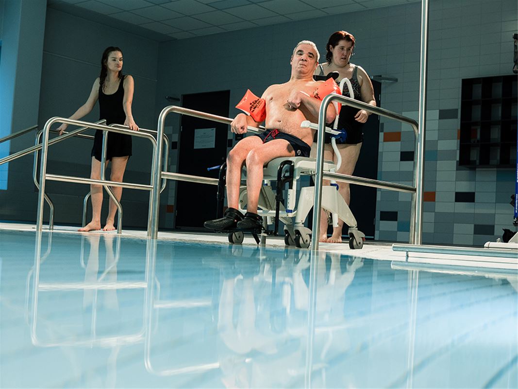 En man i rullstol som är på väg ner i en bassäng, två kvinnor i  hans närhet.