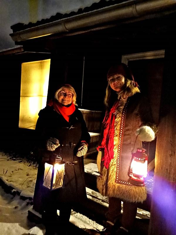 Två kvinnor som står på en trapp och håller i varsin lykta, båda har vinterkläder, det är mörkt,  en byggnad i bakgrunden, snö på marken.