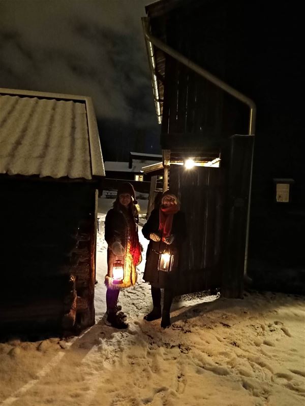 Två kvinnor i vinterkläder med varsin lykta,  de står i en öppning  in till en gård, träbyggnader, snö på marken, bilden tagen på kvällen.