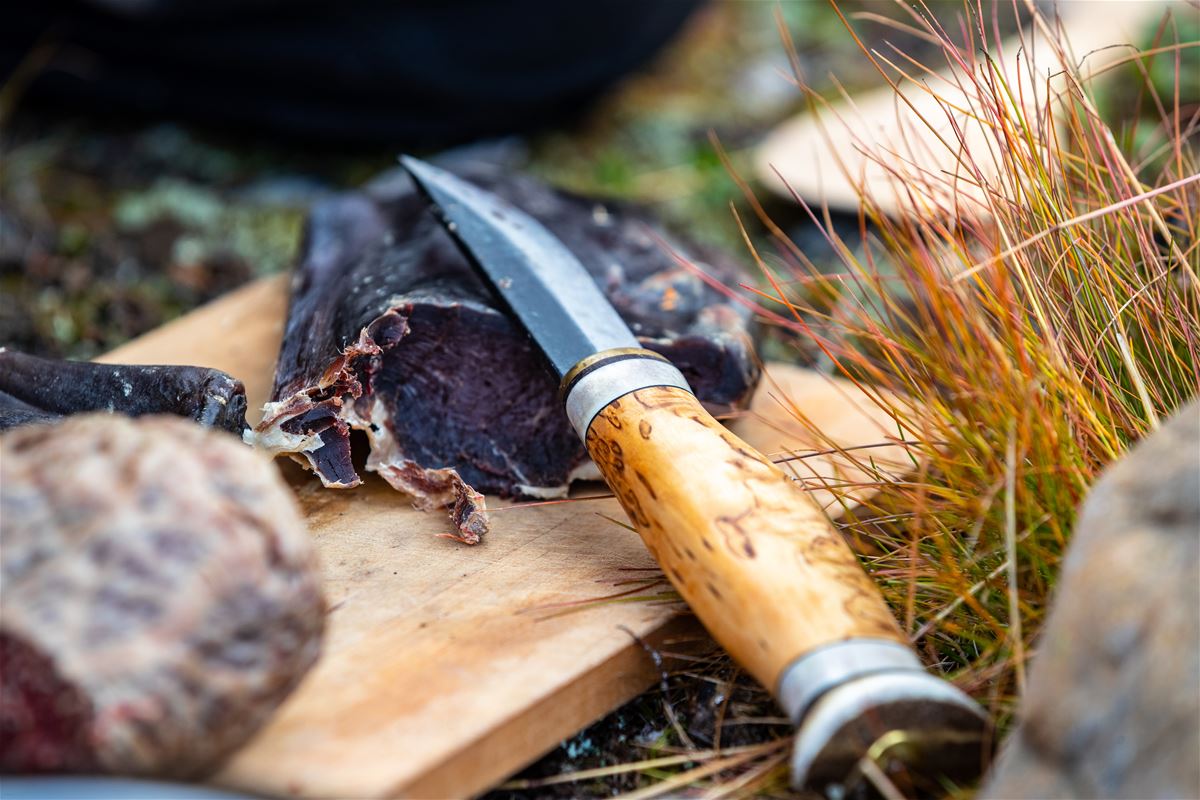 En köttbit på en skärbräda, en kniv som liggerpå skärbrädan, skärbrädan ligger i naturen, gräs syns.