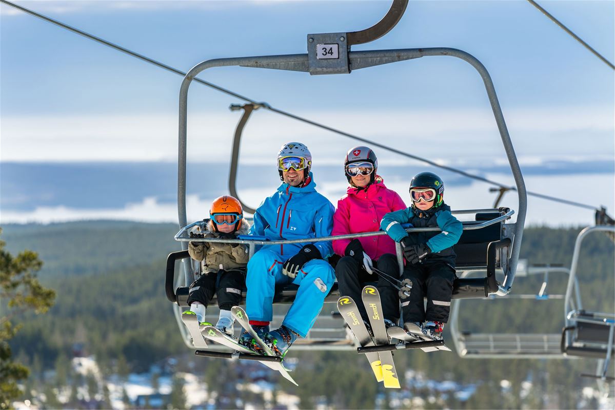 Four people in a ski lift, forest, mountains and a lake in the background.