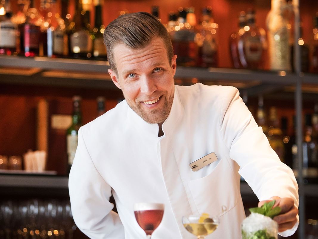 A happy waiter in a white service jacket, two wine glasses in front of him.
