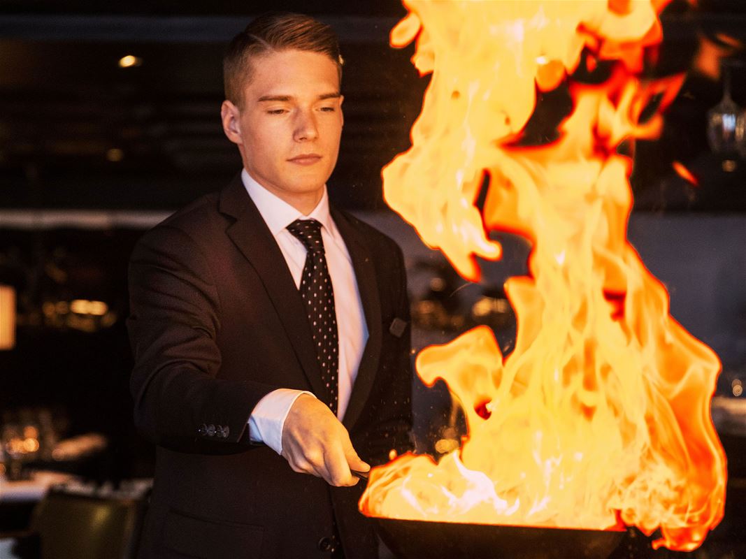 En man i svart kavaj, vit skjorta och slips håller i en stekpanna med öppen eld. 