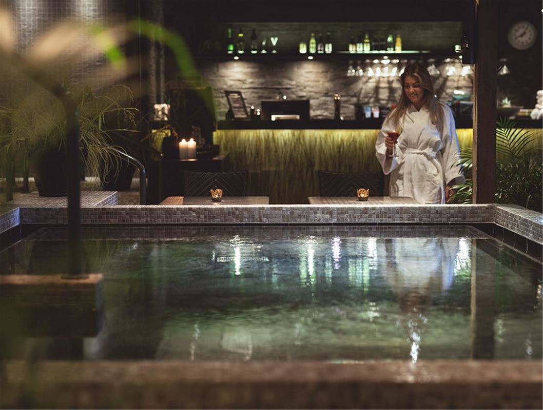 En kvinna i vit badrock som står vid en bassäng, en bar i bakgrunden.