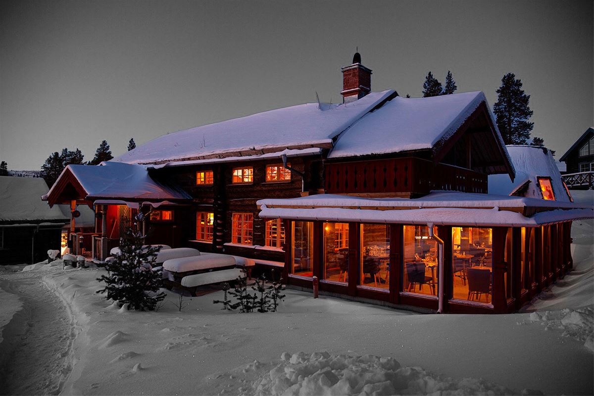 Stor träbyggnad i snölandskap med många ljus inomhus.