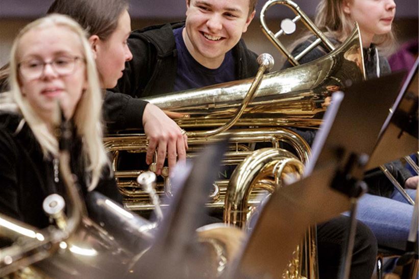 Bild på glada ungdomar som sitter och pratar med sina instrument i knät. På bilden ser vi tuba, saxofon och trombon.