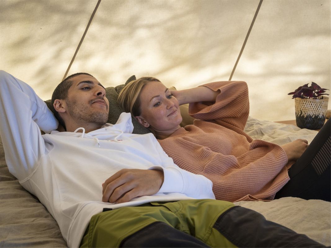 Ett par ligger i en säng intill ett glampingtält.
