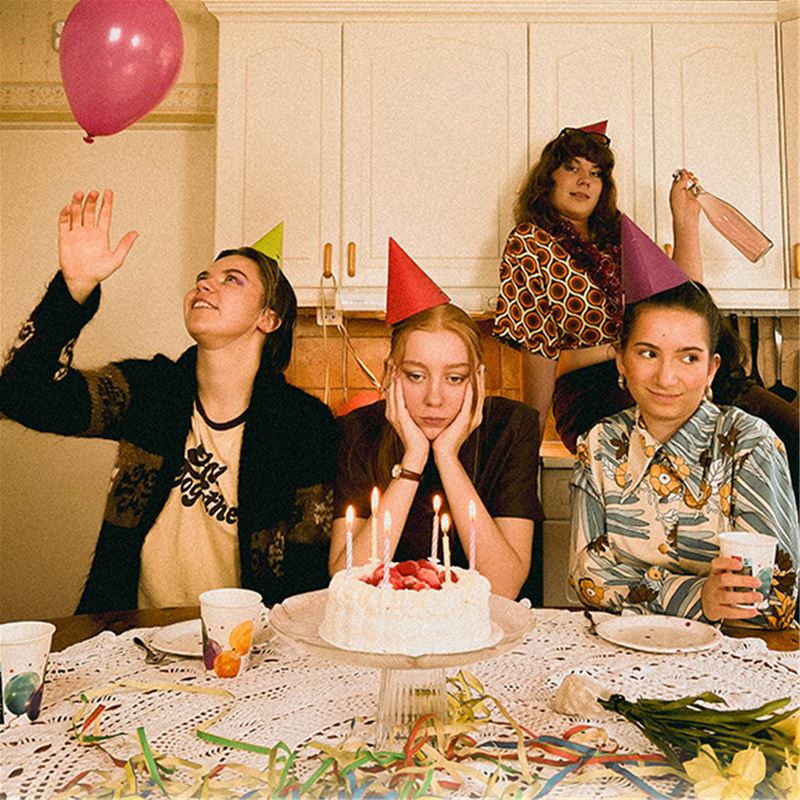 Fyra ungdomstjejer i ett kök. De verkar ha kalas, tårta med ljus på bordet, partyhattar, ballonger. Ena av tjejerna har händerna i ansiktet och verkar trött och ledsen.