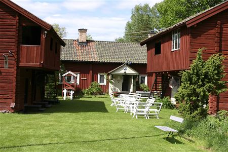 Gårdstun med röda timmerbyggnader i äldre stil, vita trädgårdsmöbler utplacerade på gräset.