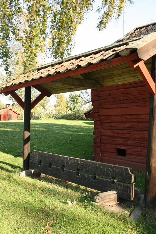 Part of a red timber building, in front of it a board with six holes, lawn and in the background red timber buildings.