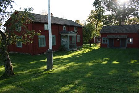 Gräsmatta, nedre delen av en flaggstång, två röda timmerbyggnader, en större i två våningar med farstukvist och en mindre.