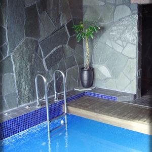 Pool med blå mosaik längs kanten, naturfärgat klinkers på golvet och väggar av grå natursten. 
