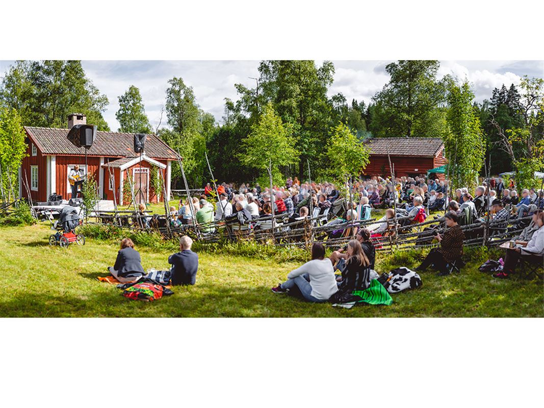 Publik sittandes i gräset framför Luossastugan där uppträdande sker framför stugan.