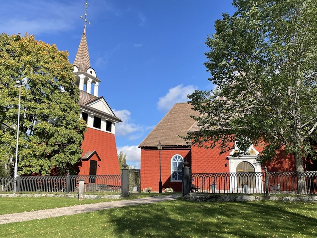 Sundborns kyrka, exteriörbild, röd träkyrka, svart järnstaket, gång fram till kyrkan, klockstapel till vänster.